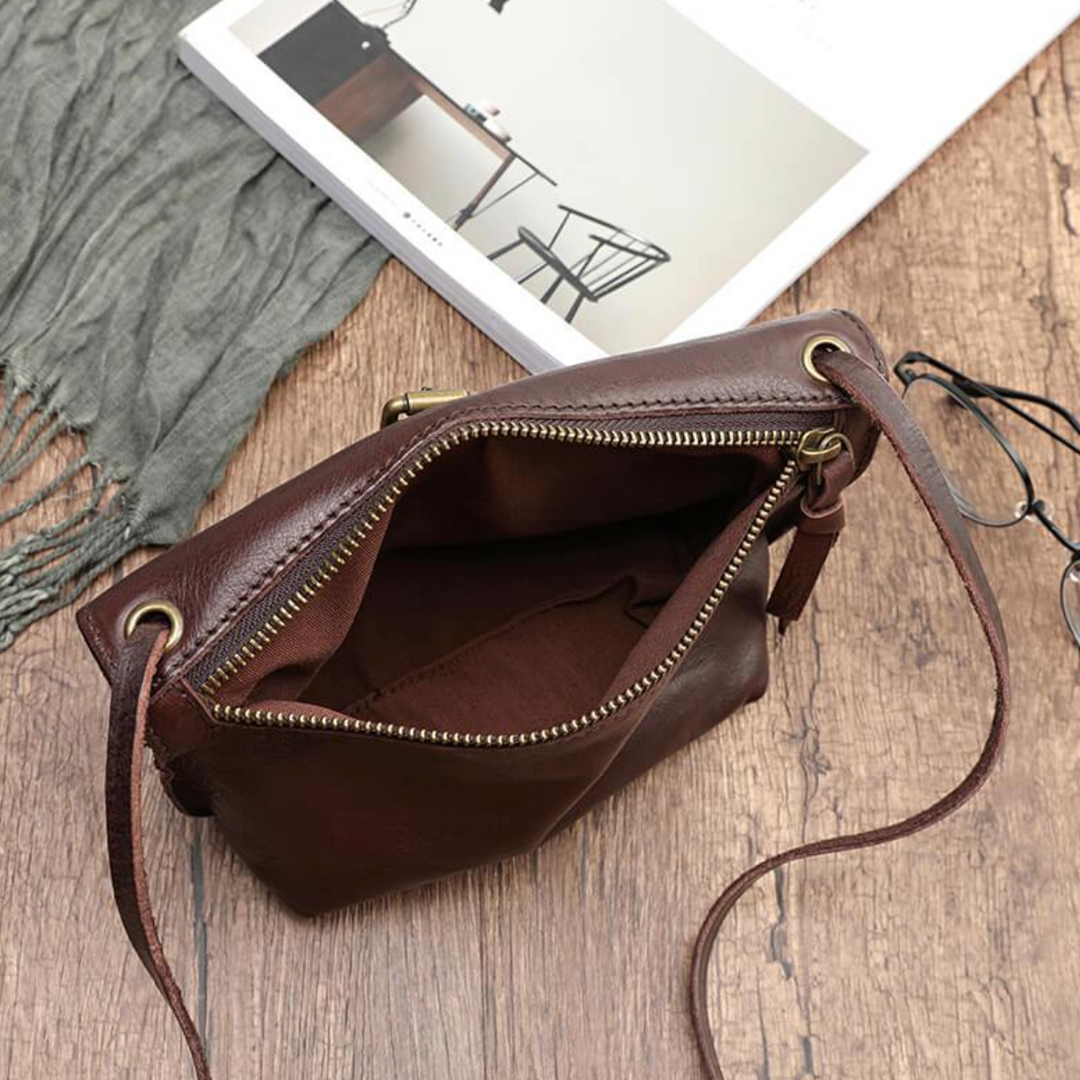 Borsa a tracolla in pelle marrone con cerniera aperta su tavolo in legno, accanto a occhiali e rivista. Elegante accessorio moda donna.