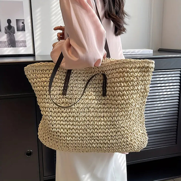 Donna con borsa di paglia intrecciata grande, manici in pelle marrone, indossa camicia rosa e gonna bianca. Accessorio moda estivo elegante.