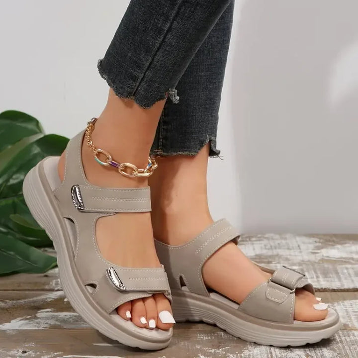 Sandali beige da donna con cinturino regolabile, suola antiscivolo e tacco basso, indossati con jeans scuri. Accessorio alla caviglia dorato.