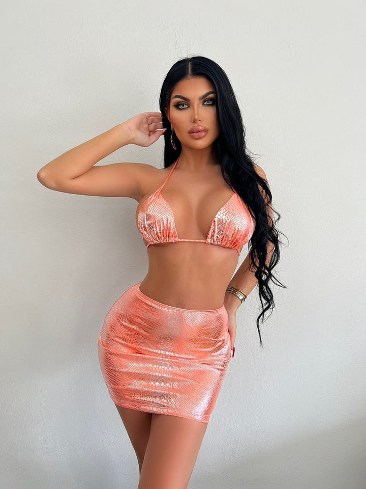 Donna in posa con top bikini e minigonna rosa scintillante, capelli lunghi e neri, sfondo bianco. Moda estiva, abbigliamento glamour.