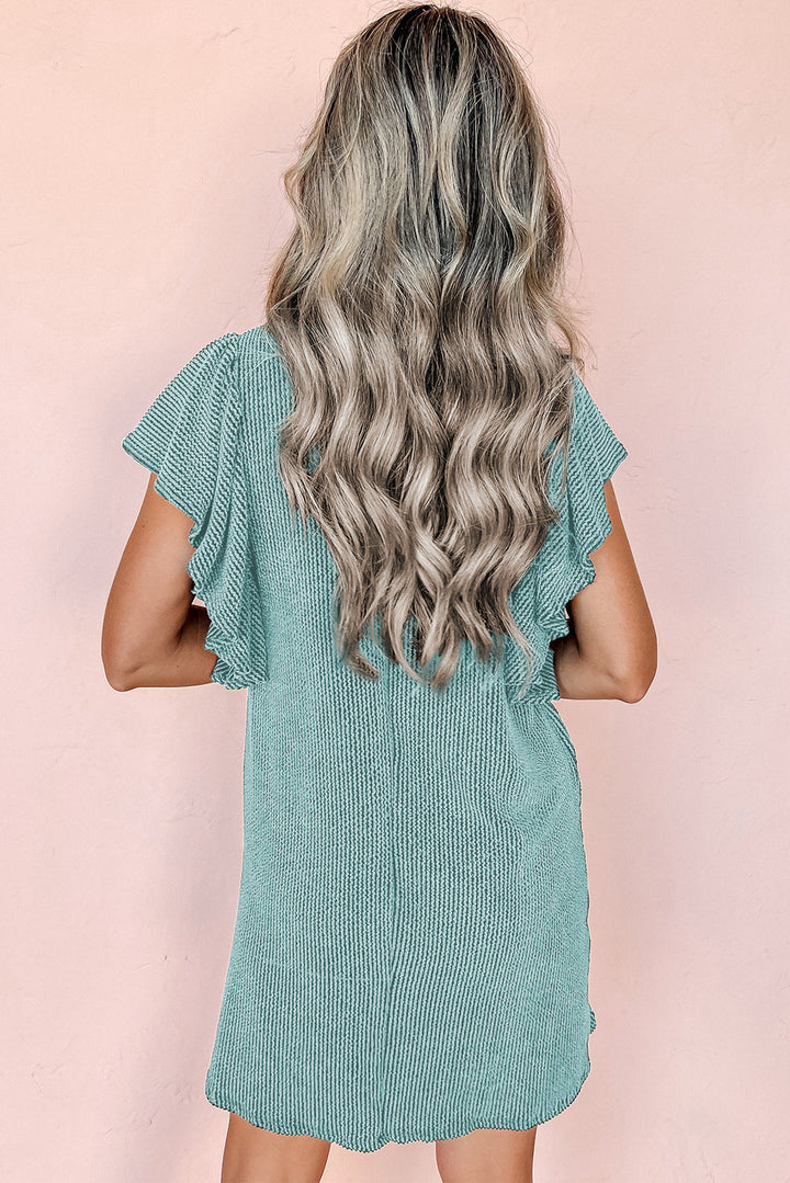 Donna con capelli ondulati biondi indossa un abito verde menta a coste con maniche corte volant, sfondo rosa. Moda estiva, abbigliamento casual.