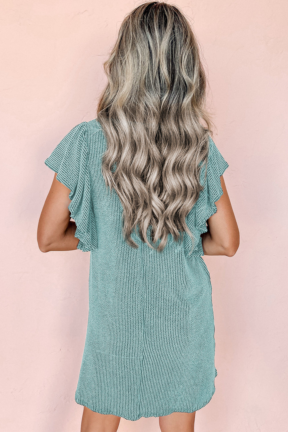 Donna con capelli ondulati biondi indossa un abito verde menta a maniche corte, sfondo rosa. Moda estiva, abbigliamento casual elegante.