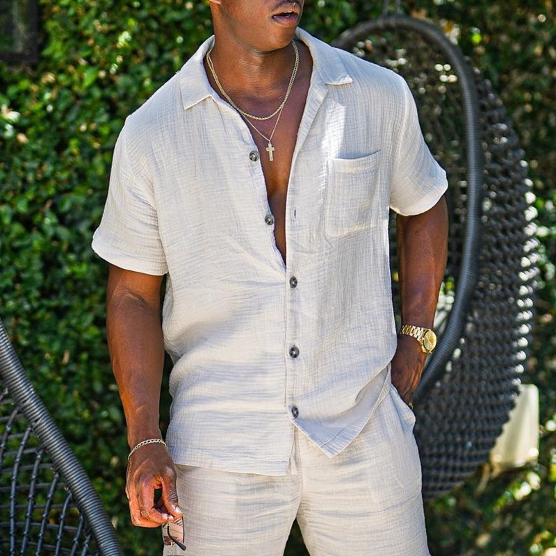Uomo con camicia di lino beige, pantaloni coordinati, collana con croce, orologio dorato, sfondo di foglie verdi. Moda estiva elegante e casual.