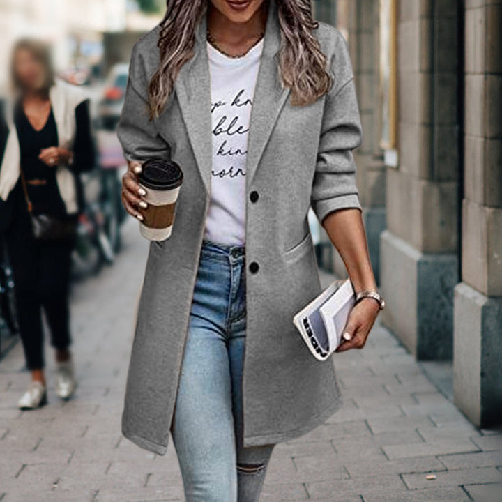 Donna con cappotto grigio elegante, jeans aderenti e maglietta bianca, tiene caffè e rivista. Moda autunnale urbana, look casual chic.