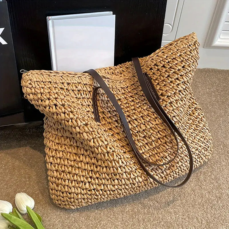 Borsa in paglia intrecciata con manici in pelle marrone, posizionata su un tappeto beige, accanto a un libro e fiori bianchi. Elegante e spaziosa.