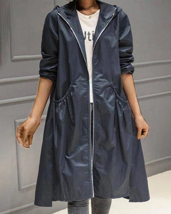 Giacca lunga impermeabile blu scuro con cappuccio, zip frontale e tasche laterali, indossata su maglietta bianca e jeans. Moda casual donna.