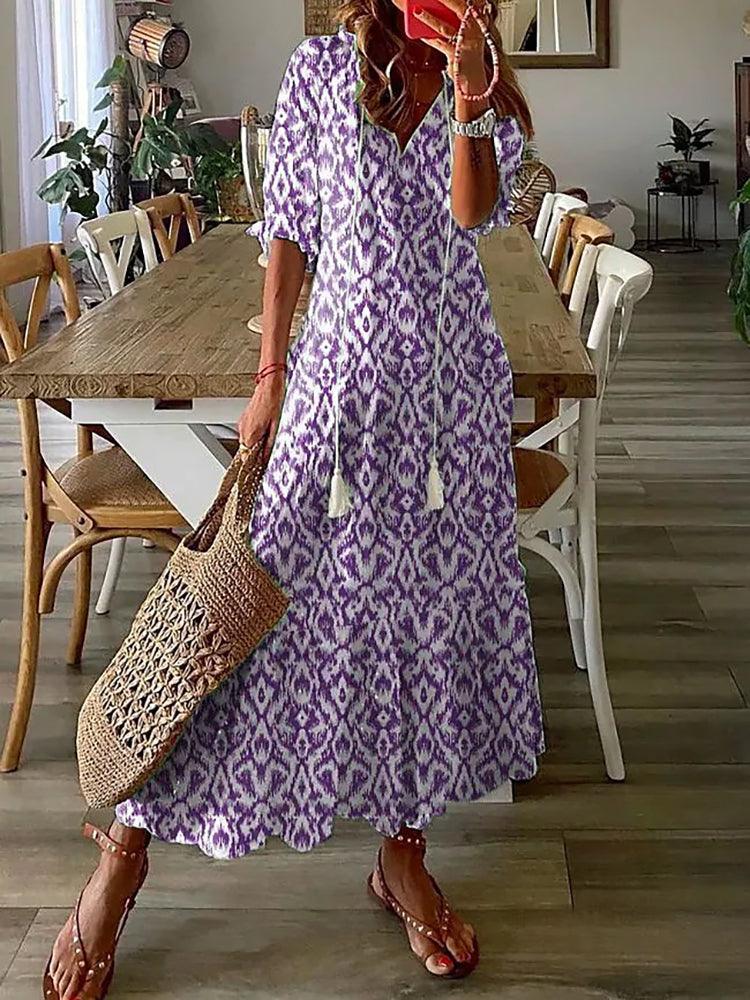 Donna con abito lungo boho viola e bianco, motivo etnico, in piedi in sala da pranzo. Borsa di paglia intrecciata, sandali marroni. Arredamento moderno.