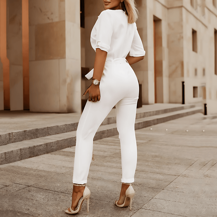 Donna in elegante completo bianco, pantaloni e camicia, tacchi alti beige, posa all'aperto su sfondo urbano. Moda femminile, abbigliamento chic.