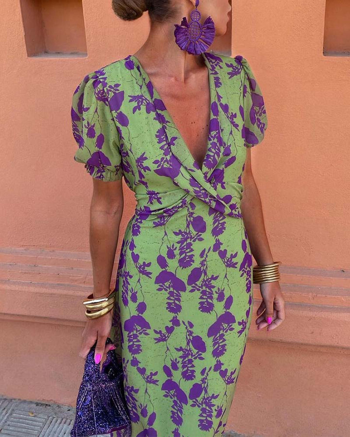 Abito verde con stampa floreale viola, maniche a sbuffo, scollo a V profondo. Accessori: orecchini viola, bracciali dorati, borsa glitter.