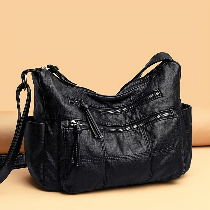 Borsa a tracolla in pelle nera con cerniere multiple su sfondo beige. Design elegante e pratico, ideale per moda donna e accessori di tendenza.