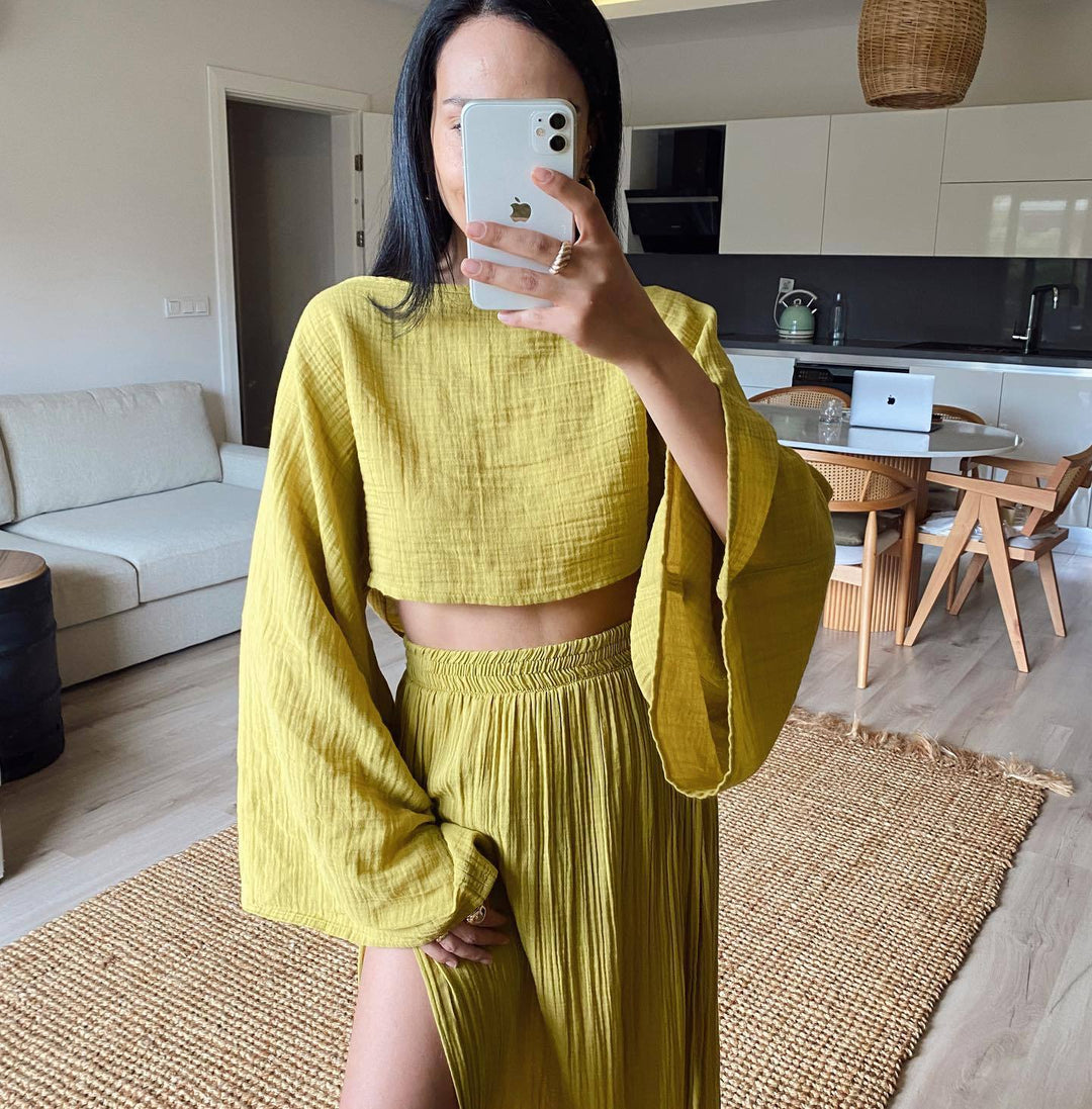 Donna con abito coordinato giallo senape, top a maniche lunghe e gonna plissettata, in soggiorno moderno. Moda estiva, abbigliamento casual elegante.