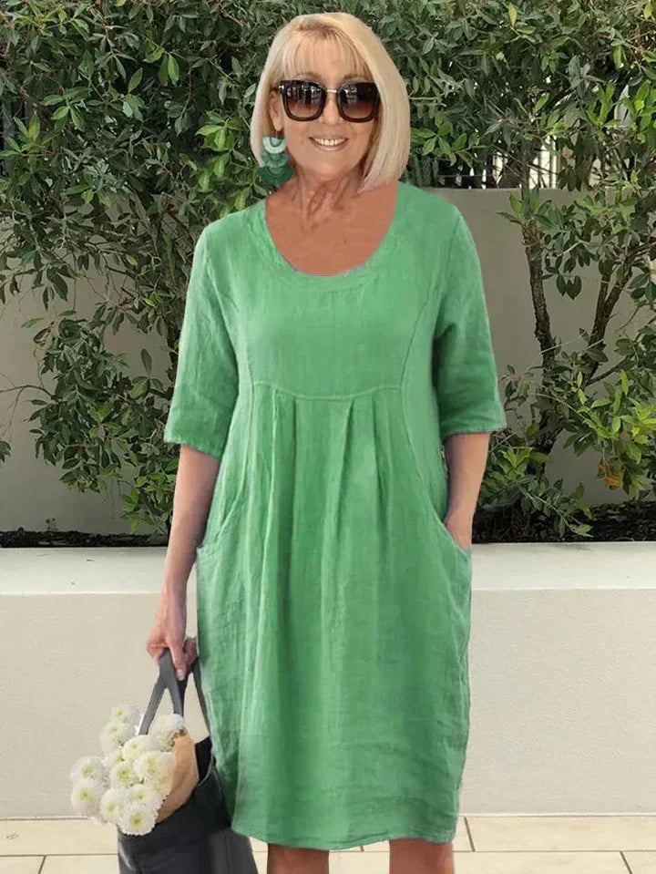 Donna con abito verde in lino, occhiali da sole, orecchini turchesi, borsa nera e fiori bianchi. Moda estiva casual, look elegante e fresco.