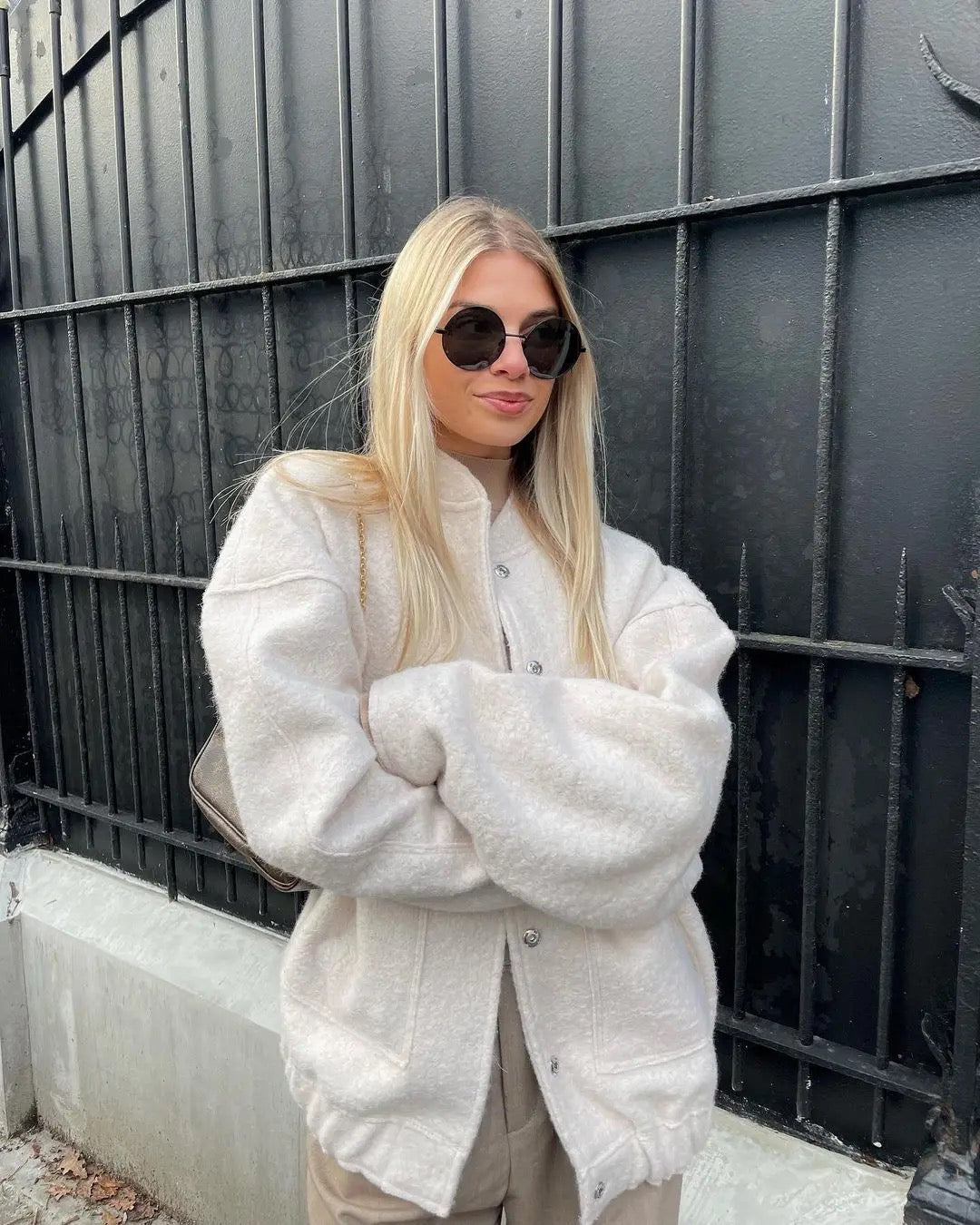 Giovane donna con occhiali da sole e giacca bianca, posa davanti a una recinzione nera. Moda autunnale, stile casual chic, look urbano.