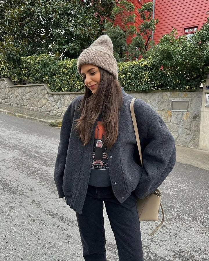 Donna con cappotto grigio e berretto beige, in piedi su strada urbana. Sfondo con muro di pietra e vegetazione. Moda invernale casual.
