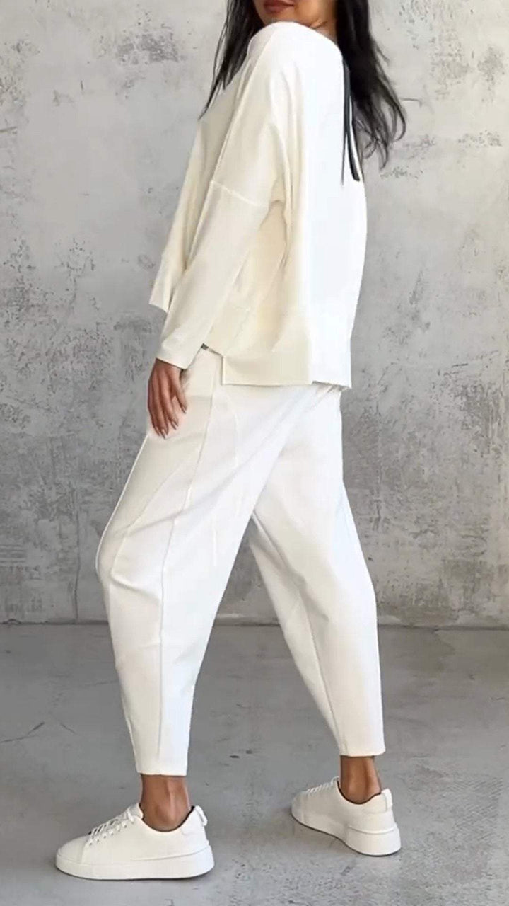 Donna in abbigliamento casual bianco, indossa maglia e pantaloni larghi, scarpe da ginnastica bianche, posa su sfondo grigio. Moda minimalista elegante.