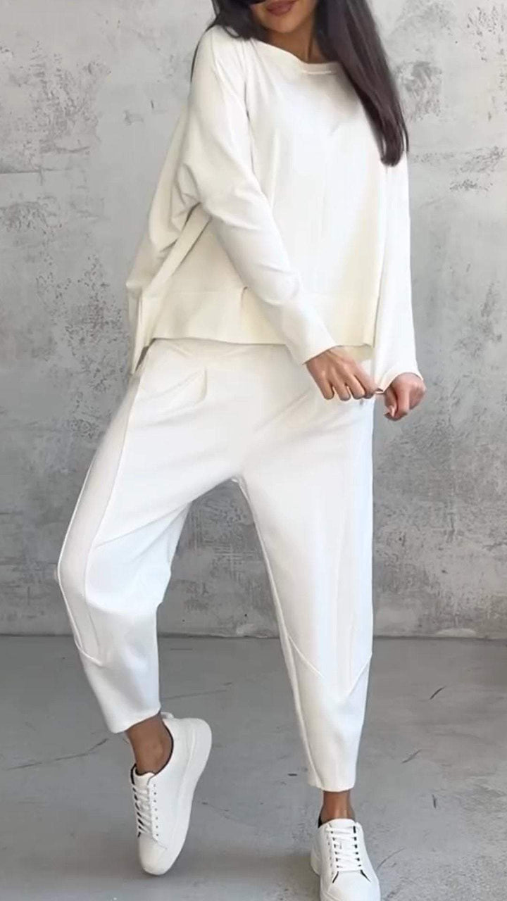 Donna in completo bianco casual, composto da maglia e pantaloni larghi, con sneakers bianche. Look minimalista e moderno su sfondo grigio.