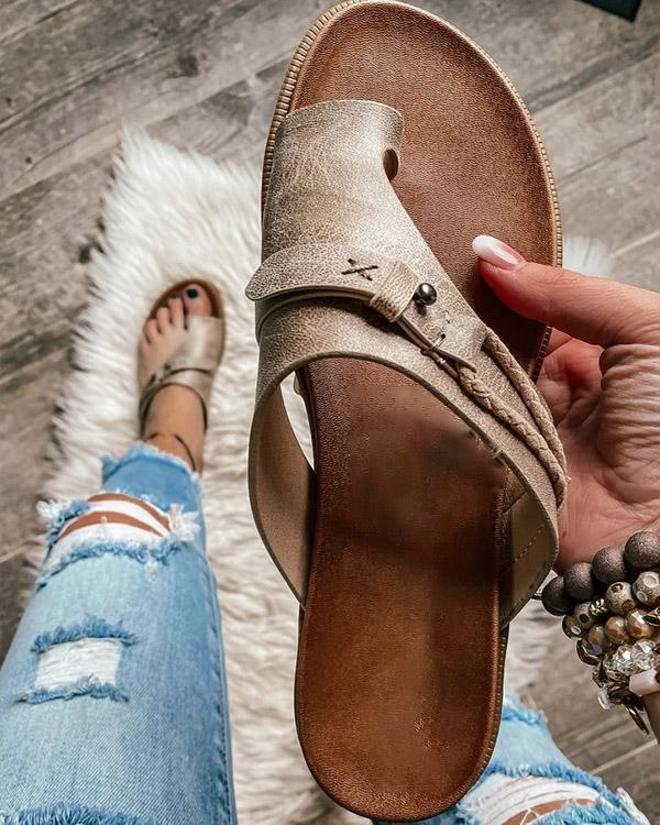 Sandali in pelle marrone chiaro con dettagli intrecciati, tenuti in mano su sfondo di pavimento in legno e tappeto bianco, jeans strappati visibili.