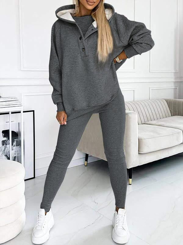 Donna in tuta sportiva grigia, felpa con cappuccio e pantaloni aderenti, scarpe da ginnastica bianche, in salotto moderno. Abbigliamento casual elegante.