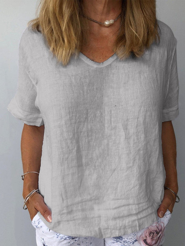 Donna indossa maglietta di lino grigia con scollo a V, abbinata a pantaloni bianchi floreali. Moda estiva casual, abbigliamento donna elegante.