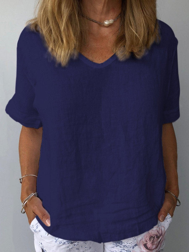Donna con maglietta blu scuro in lino, scollo a V, maniche corte, abbinata a pantaloni bianchi con stampa floreale. Moda estiva casual elegante.