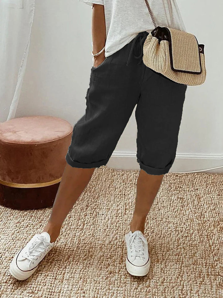 Pantaloncini neri casual da donna, abbinati a maglietta bianca e borsa a tracolla beige, su tappeto intrecciato. Scarpe da ginnastica bianche.