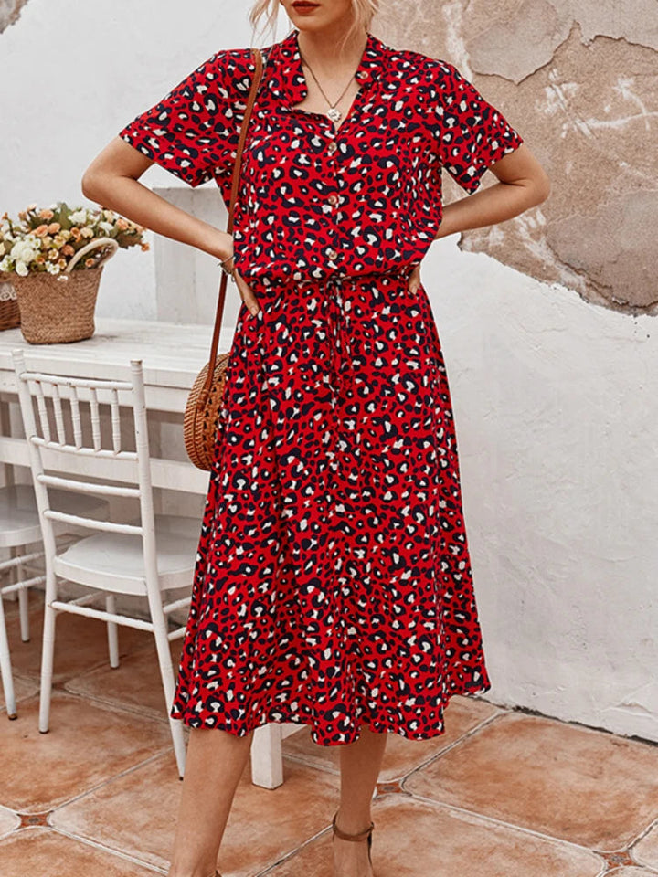 Abito rosso a fantasia leopardata, maniche corte, lunghezza media, indossato da donna. Sfondo con tavolo bianco e cesto di fiori.