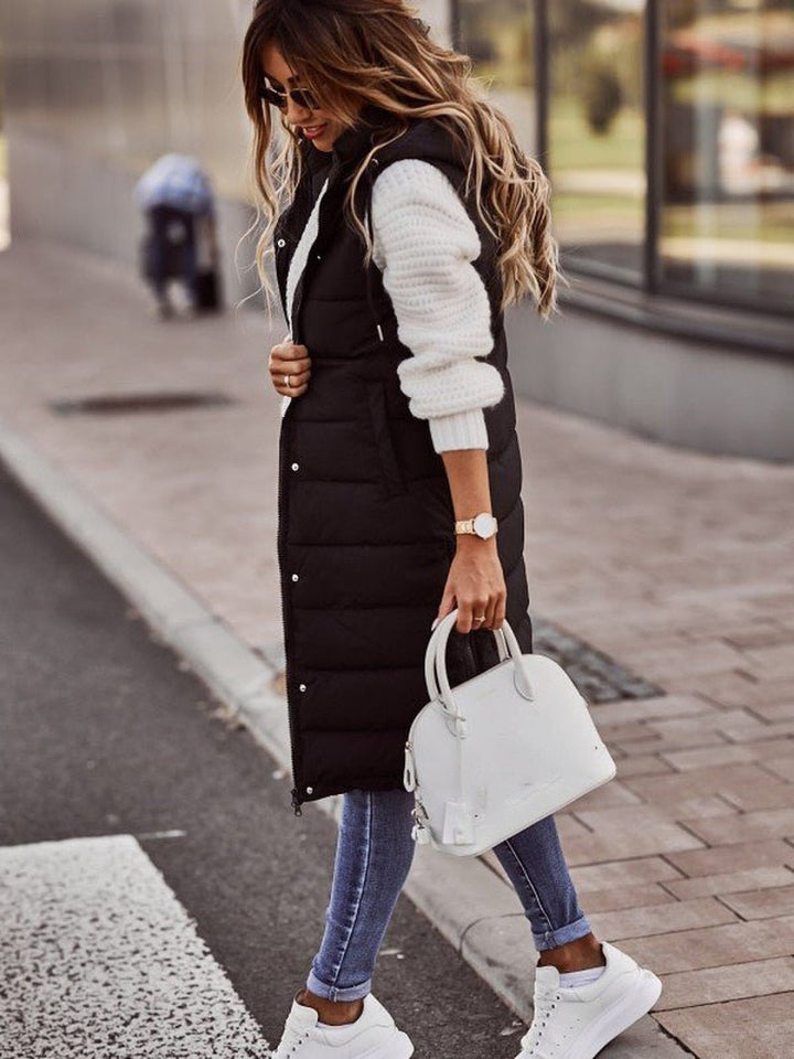 Donna con gilet lungo nero imbottito, maglione bianco, jeans aderenti e sneakers bianche, borsa bianca. Moda autunnale casual chic.