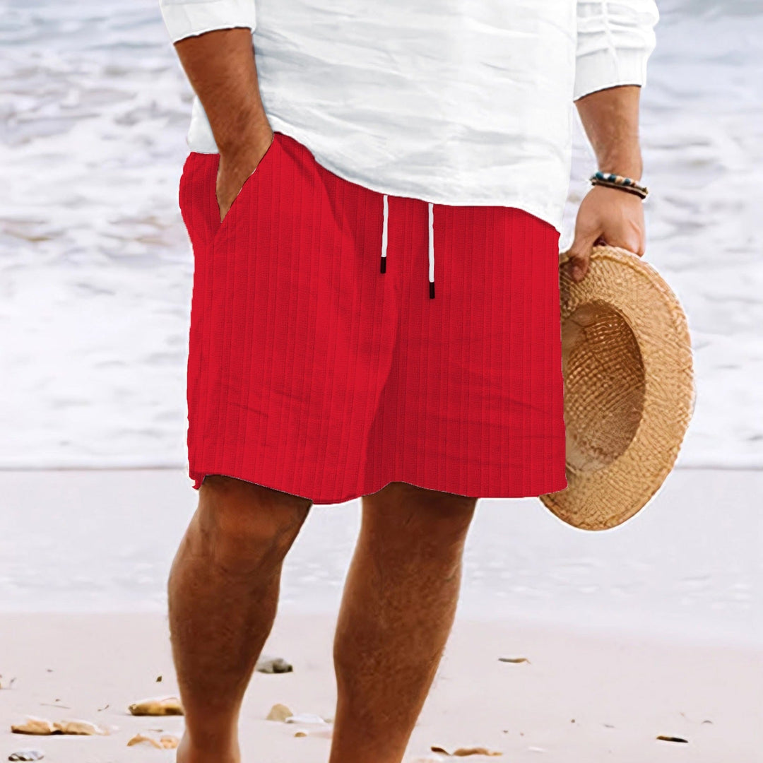 Uomo in spiaggia con pantaloncini rossi e camicia bianca, tiene un cappello di paglia. Moda estiva, abbigliamento casual mare.