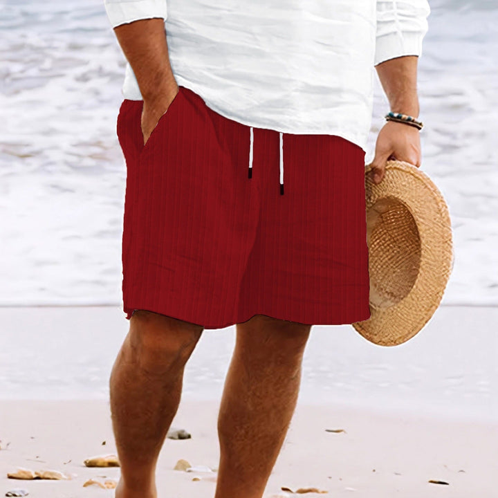 Uomo in spiaggia con pantaloncini rossi e camicia bianca, tiene cappello di paglia. Moda estiva, abbigliamento casual mare.