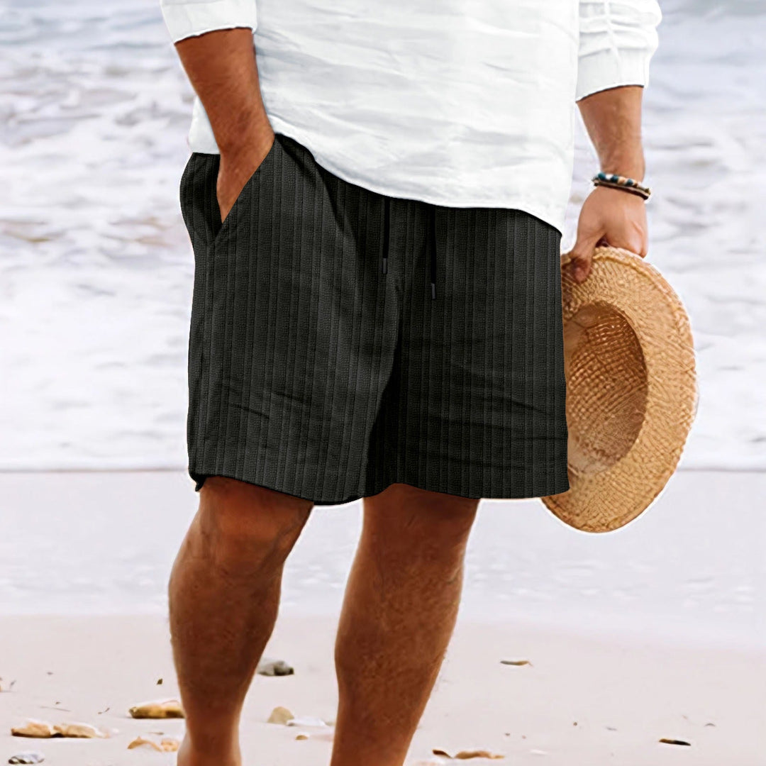 Uomo in pantaloncini neri a righe su spiaggia, indossa camicia bianca e tiene cappello di paglia. Moda estiva, abbigliamento casual mare.