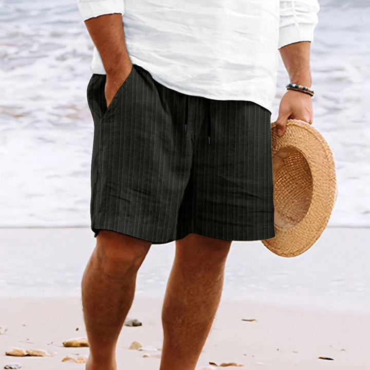 Uomo in pantaloncini neri a righe su spiaggia, indossa camicia bianca e tiene cappello di paglia. Moda estiva, abbigliamento casual mare.