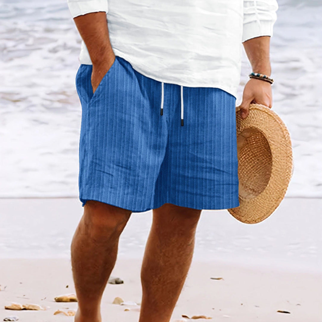 Uomo in spiaggia con pantaloncini blu a righe, camicia bianca e cappello di paglia. Moda estiva, abbigliamento casual mare.