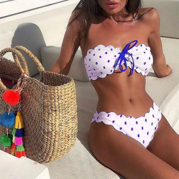 Donna in bikini bianco a pois blu su divano esterno, con borsa di paglia decorata con pompon colorati. Moda mare estiva, stile elegante.