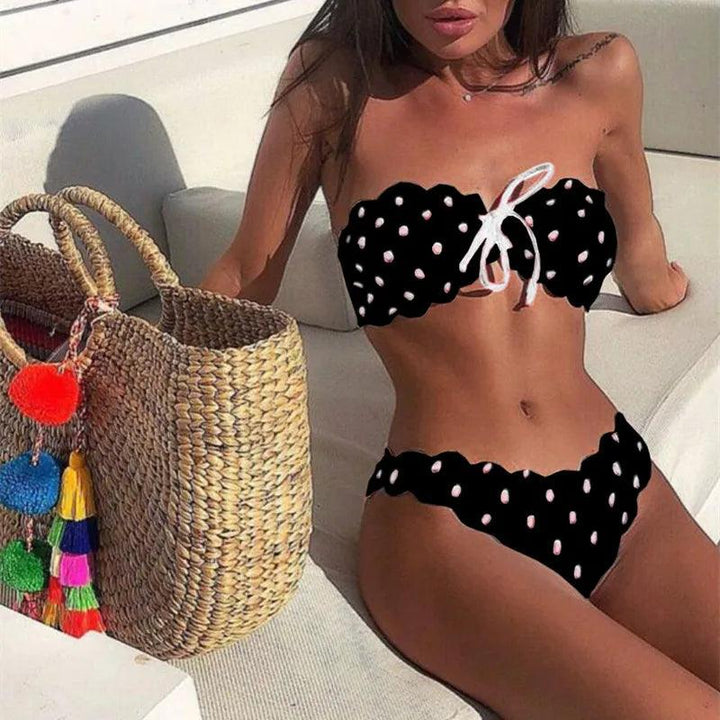 Donna in bikini nero a pois bianchi su divano bianco, con borsa di paglia decorata con pompon colorati. Moda mare estiva, stile elegante.