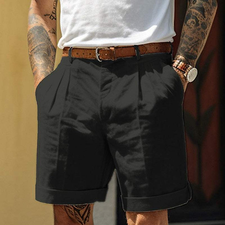 Uomo con pantaloncini neri eleganti, cintura marrone, camicia bianca. Look estivo casual, moda maschile, tatuaggi visibili, orologio al polso.