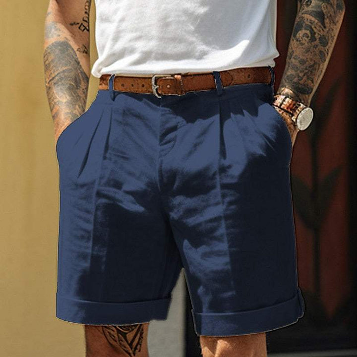 Uomo con pantaloncini blu eleganti, cintura marrone, orologio al polso e tatuaggi sulle braccia. Moda estiva maschile casual chic.